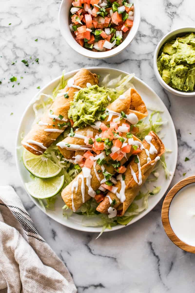 A plate of crispy taquitos topped with guacamole, pico de gallo, and a drizzle of crema, served on shredded lettuce with lime wedges. Bowls of guacamole, pico de gallo, and crema are on the side.
