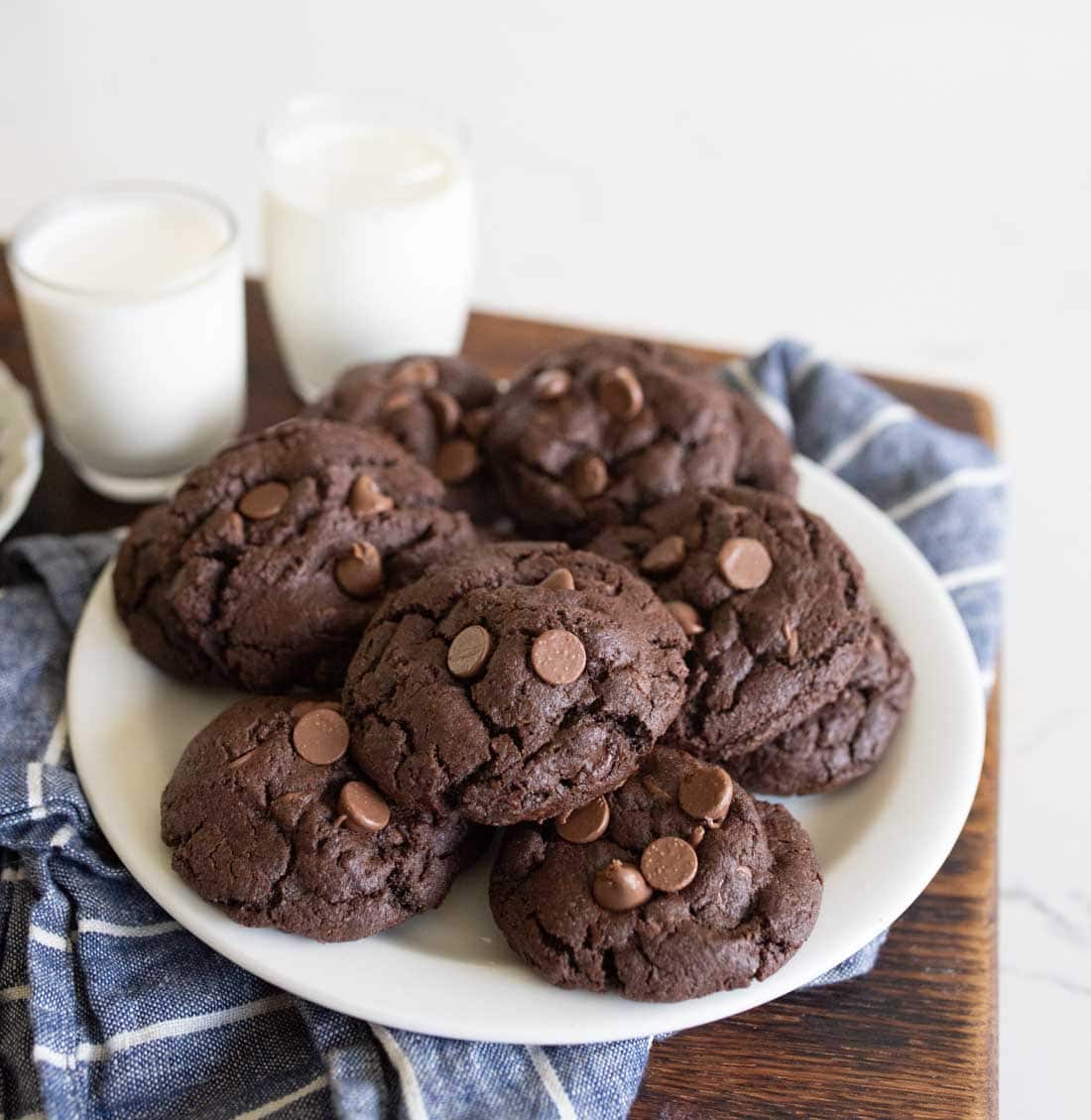 A white plate filled with chocolate chocolate-chip cookies sits on a blue and white cloth, with two glasses of milk in the background on a wooden surface.