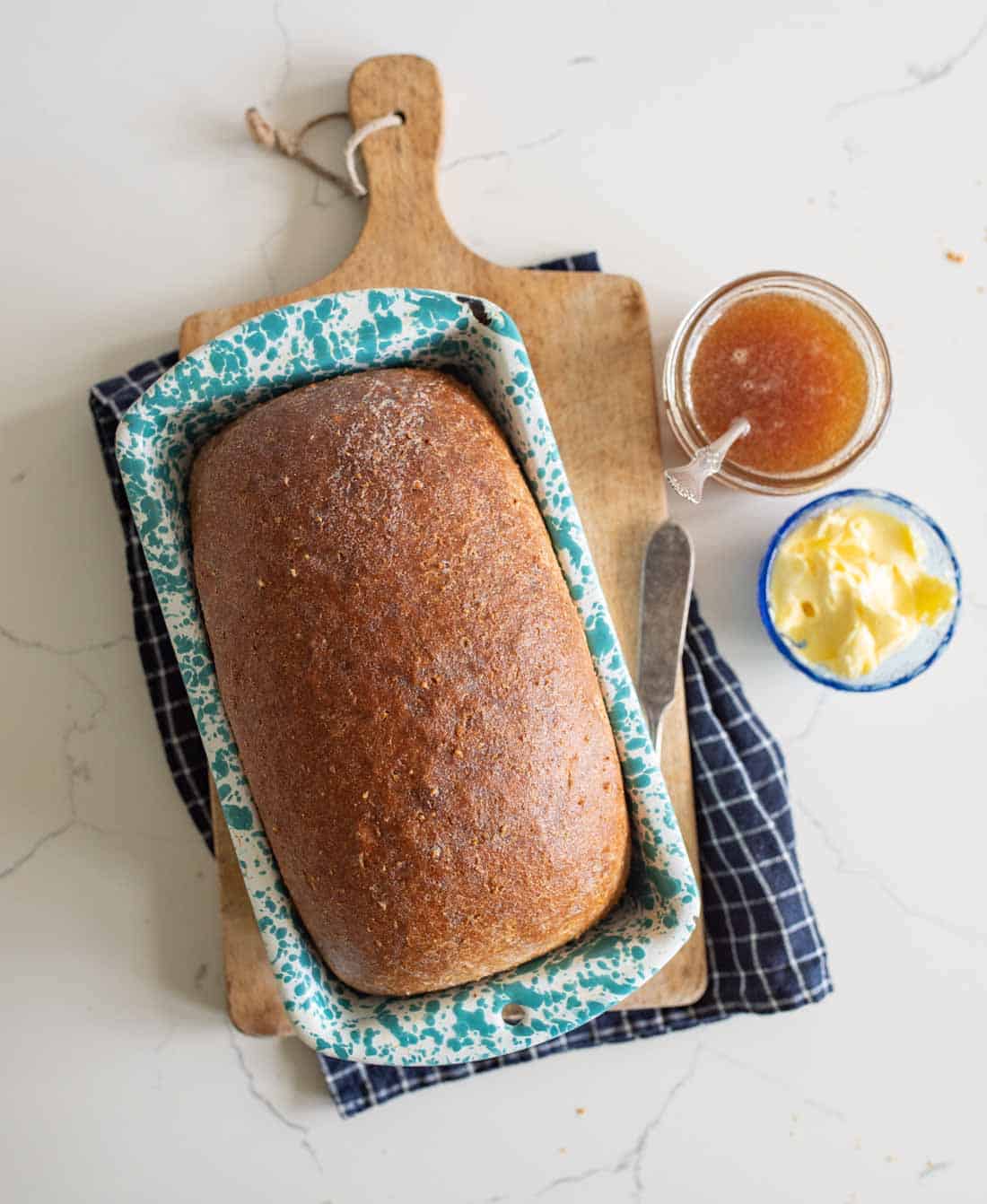 A loaf of bread in a blue speckled pan on a wooden cutting board, with a jar of jam, a dish of butter, and a knife nearby, all on a checkered cloth on a white countertop.