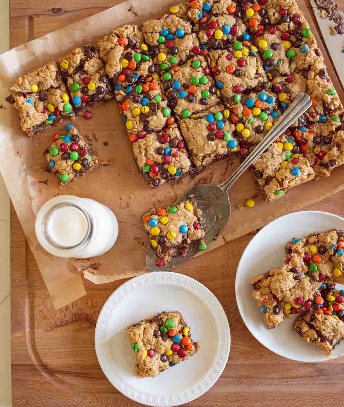 A tray of colorful M&M cookie bars on parchment paper, with squares cut and served on white plates. A metal spatula rests on the tray, and a glass of milk sits nearby on a wooden surface.