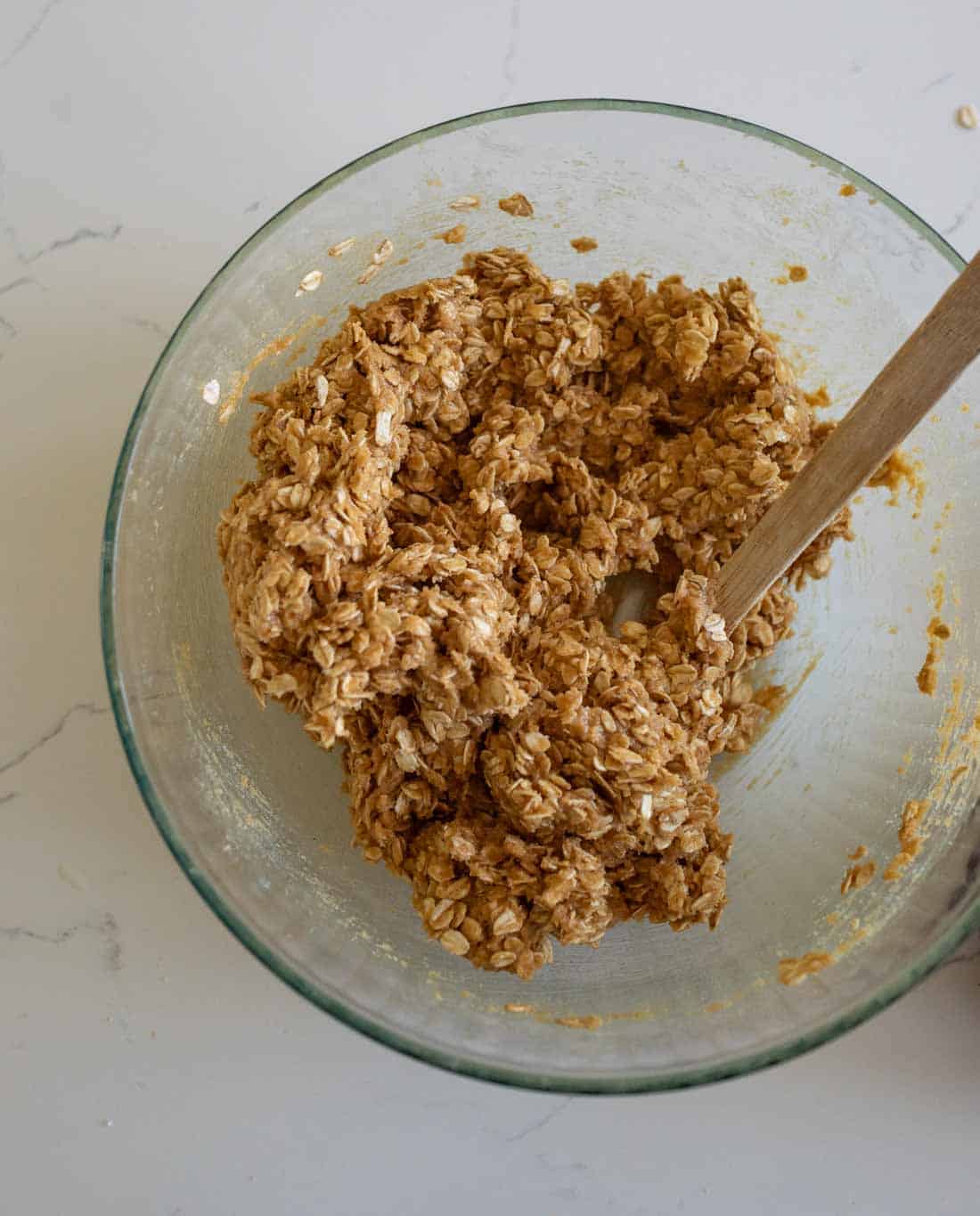 A glass bowl with a mixture of oats and a brown, sticky ingredient being stirred with a wooden spoon, sitting on a white countertop.