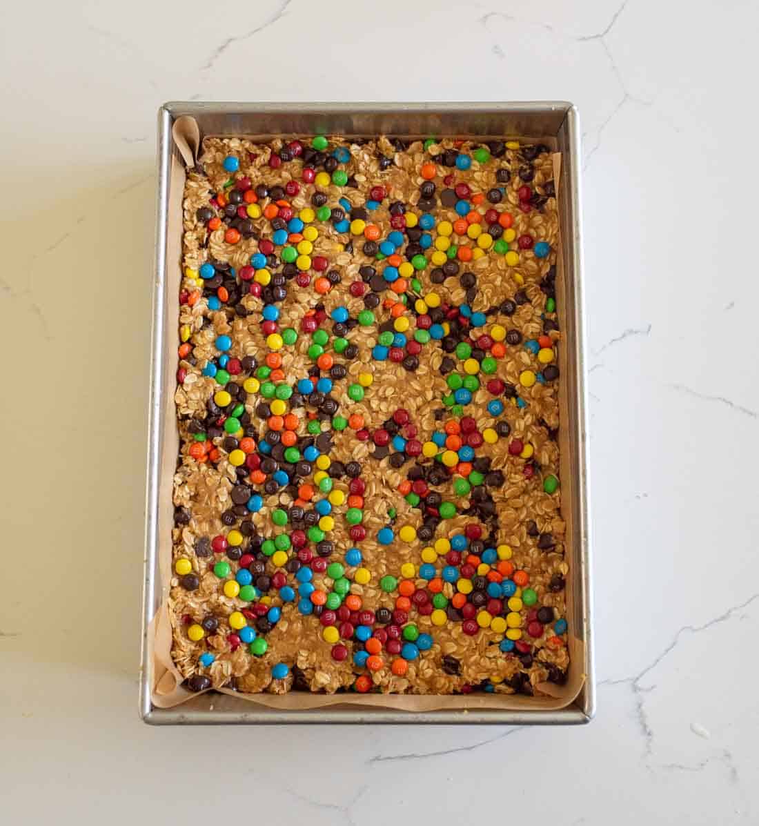A rectangular baking pan filled with a baked oat bar mixture topped with colorful chocolate candies and chocolate chips, sitting on a white marble surface.