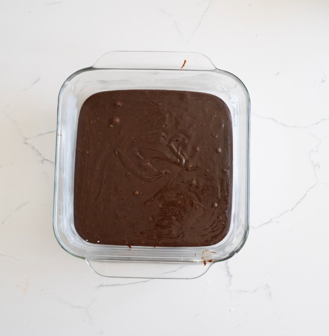 A square glass baking dish filled with chocolate brownie batter sits on a white marble countertop.