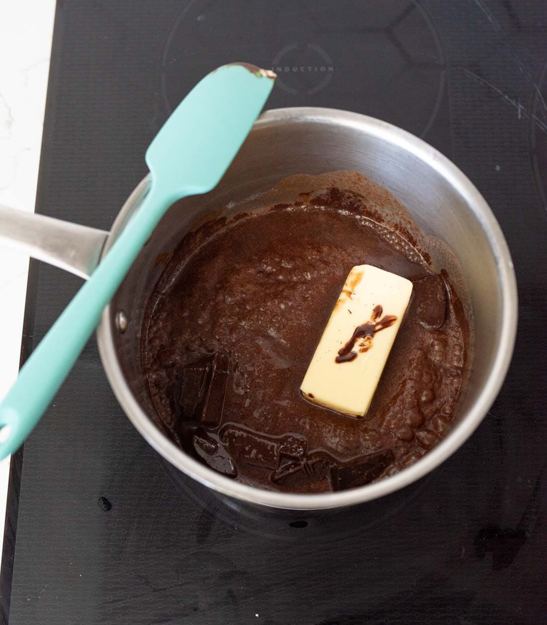 A saucepan on a stovetop contains a melted chocolate mixture with a stick of butter and some vanilla extract. A teal spatula rests on the pan’s edge.