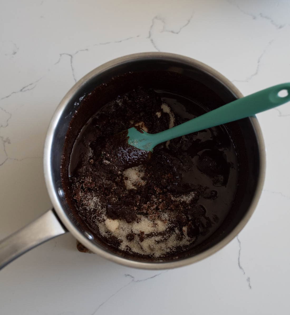 A saucepan filled with a dark chocolate mixture and sugar being stirred with a green spatula, sitting on a white marble countertop.