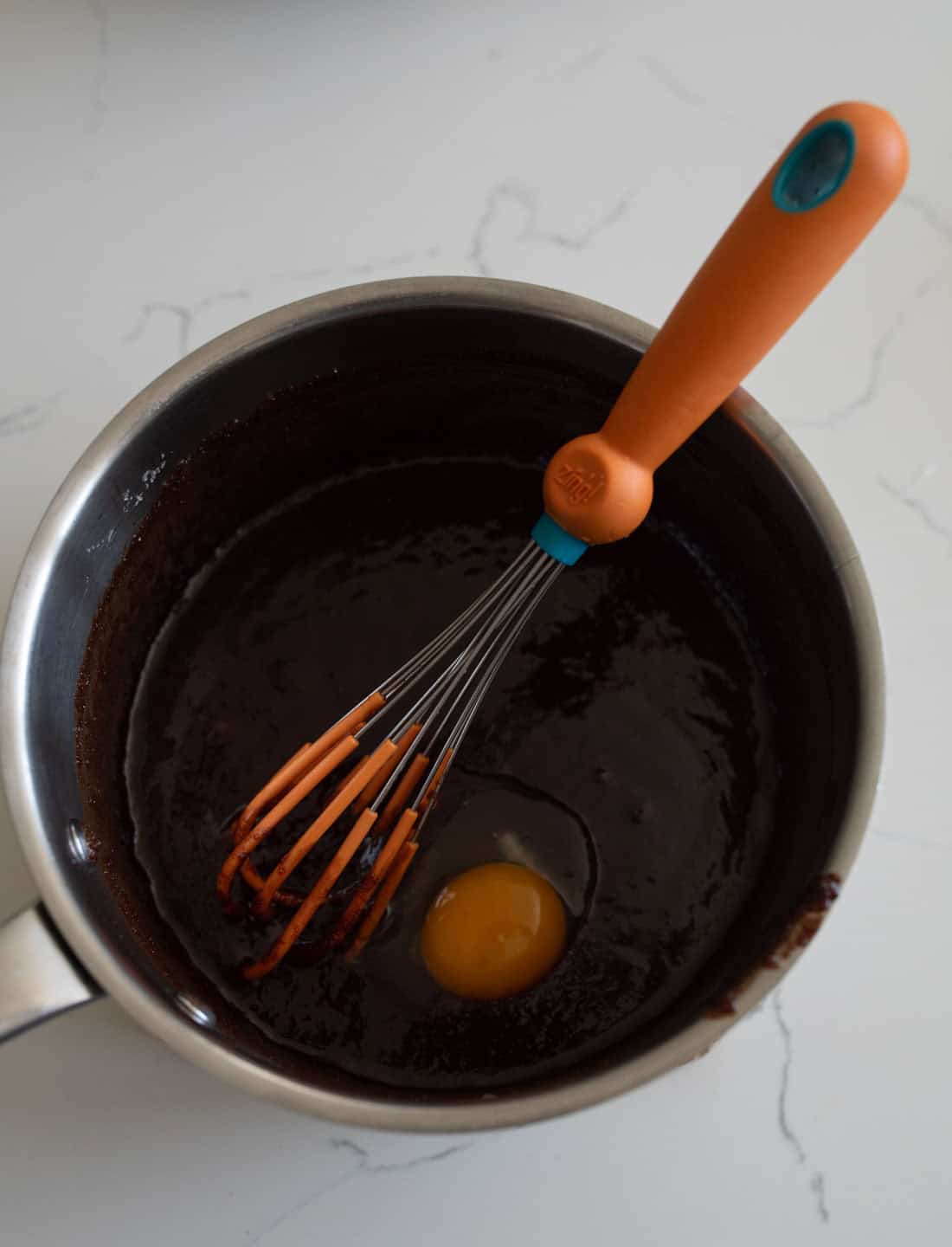 A metal saucepan containing a dark chocolate mixture with a raw egg and an orange whisk resting inside, on a white countertop.