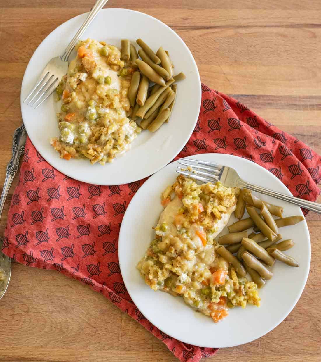 Two white plates with chicken casserole, mixed vegetables, and cooked green beans on a wooden table, with a red patterned cloth underneath. Each plate has a fork.