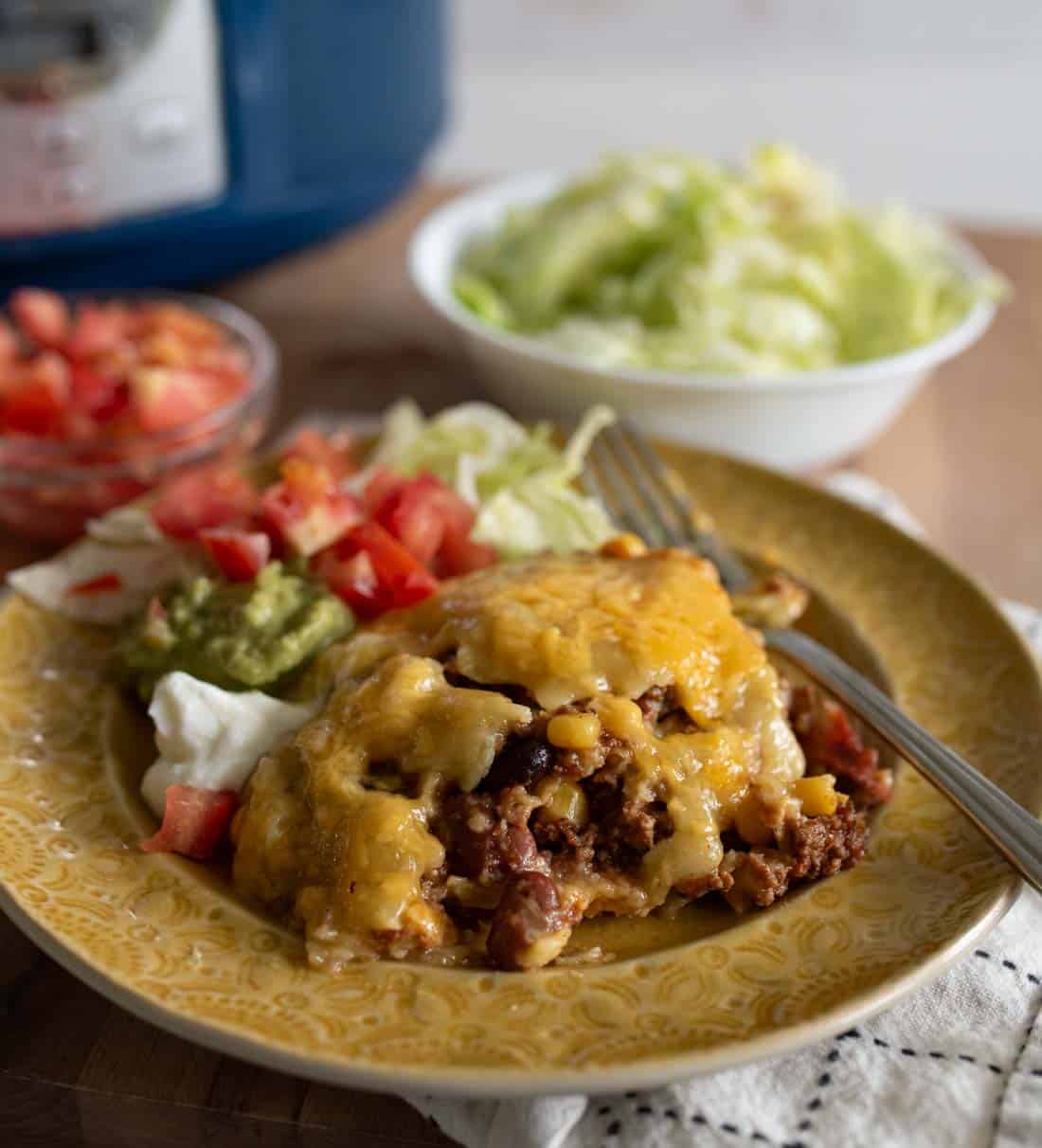 A plate with cheesy slow cooker beef enchiladas, corn, melted cheese, and sides of chopped tomatoes, guacamole, sour cream, and lettuce. A bowl of shredded lettuce and another with diced tomatoes are in the background.