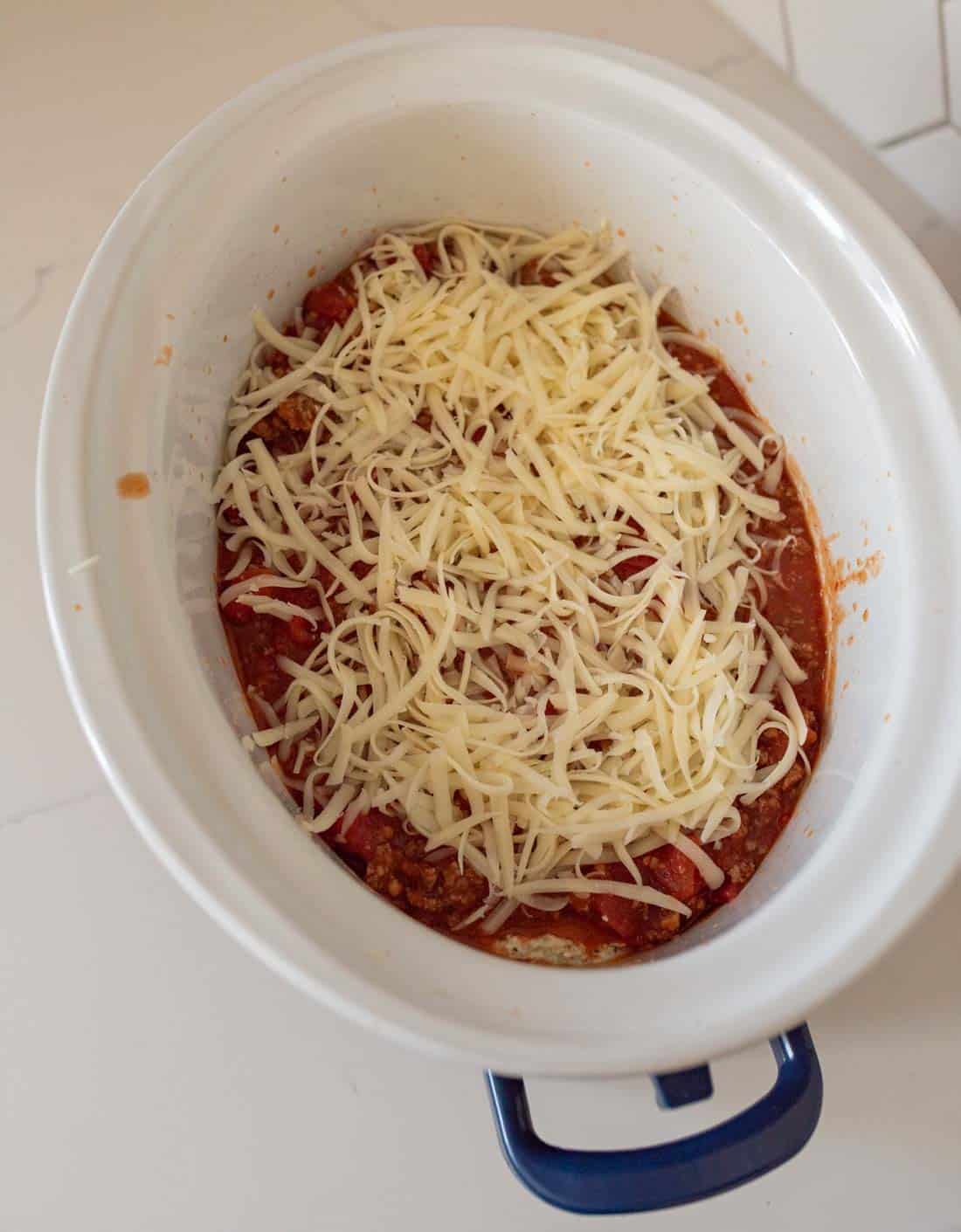 A slow cooker filled with a layer of tomato sauce, ground meat, and shredded cheese, prepared and ready to be cooked.