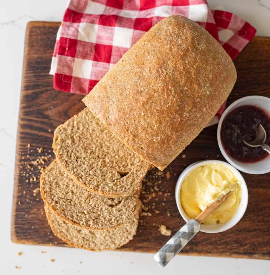 A loaf of whole wheat bread, partly sliced, sits on a wooden cutting board with a red and white checkered cloth, next to small bowls of butter and dark jam.
