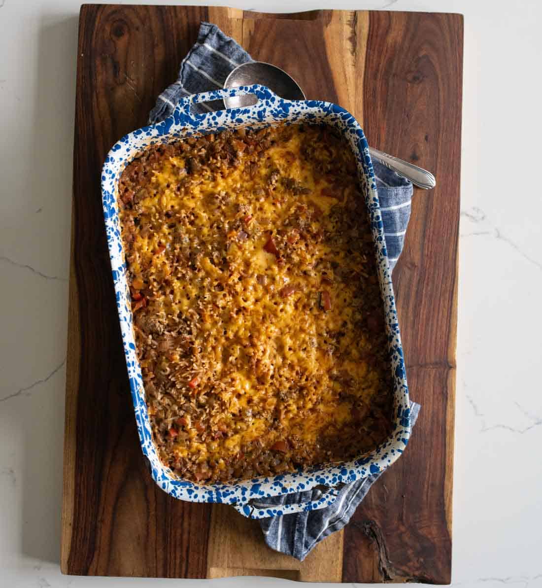 A baked casserole topped with melted cheese sits in a blue and white speckled dish on a wooden cutting board, with a blue cloth partially tucked underneath.