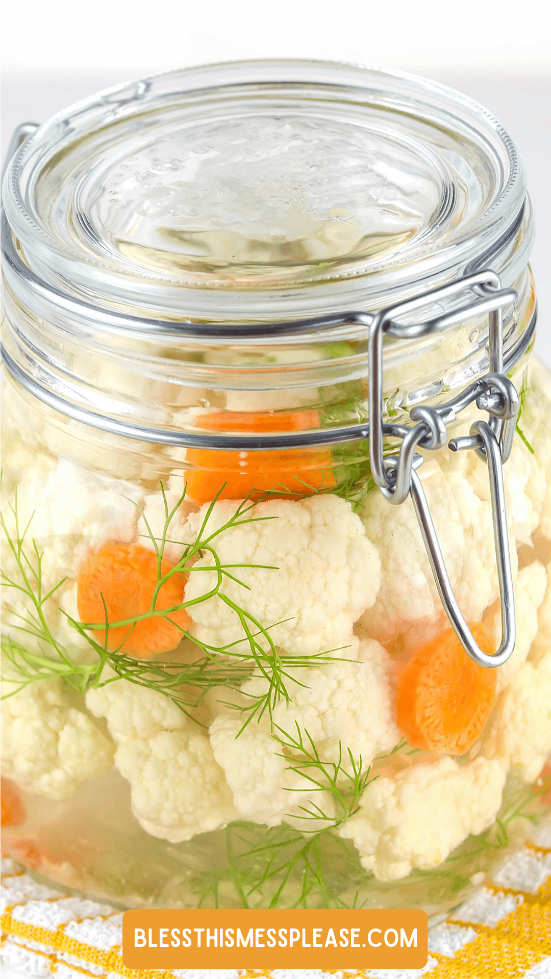 A glass jar filled with pickled cauliflower, sliced carrots, and fresh dill sprigs in brine, sealed with a metal clasp lid. Learn how to preserve cauliflower at displayed at the bottom.