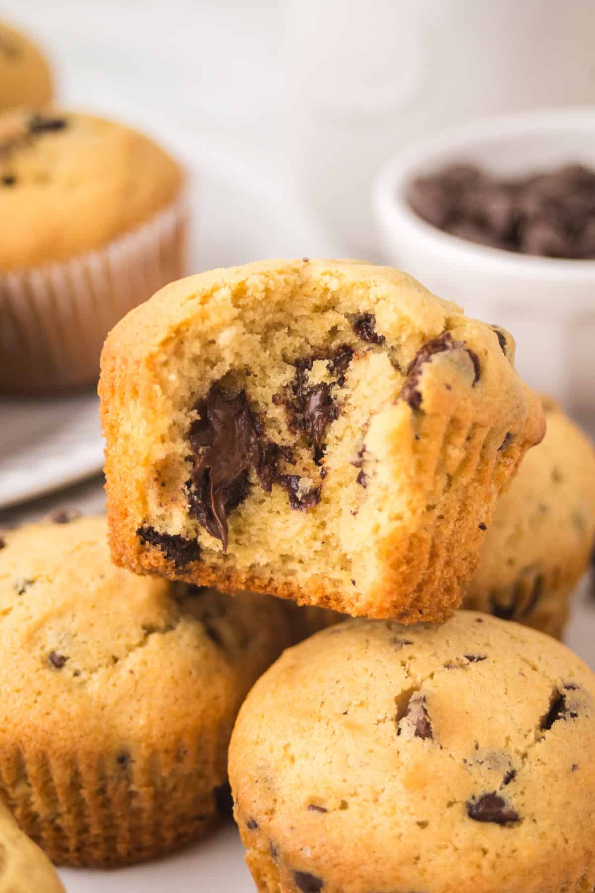 A close-up of chocolate chip muffins, with one muffin showing a bite taken out, revealing gooey melted chocolate chunks inside. Other whole muffins and a bowl of chocolate chips are in the background.