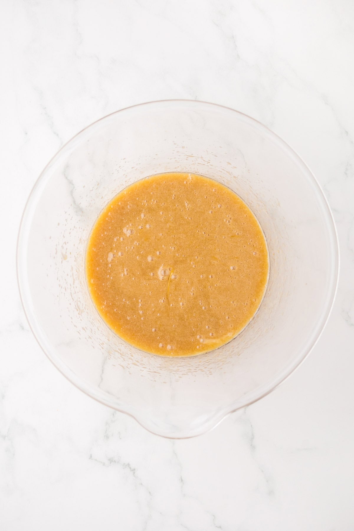 A clear glass mixing bowl with a light brown, slightly frothy liquid batter sits on a white marble surface.