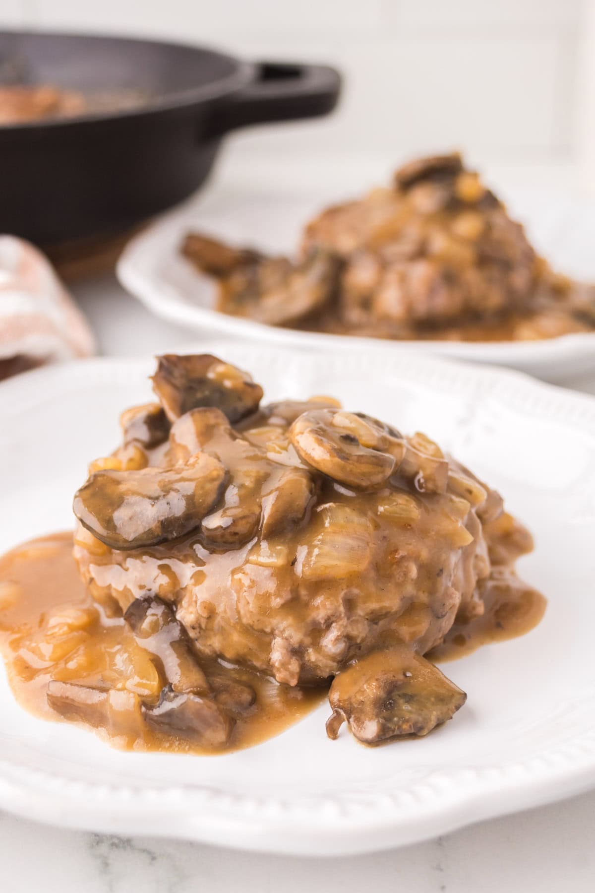 A chopped steak patty topped with brown mushroom and onion gravy is served on a white plate, with another plate of the same dish and a skillet blurred in the background.