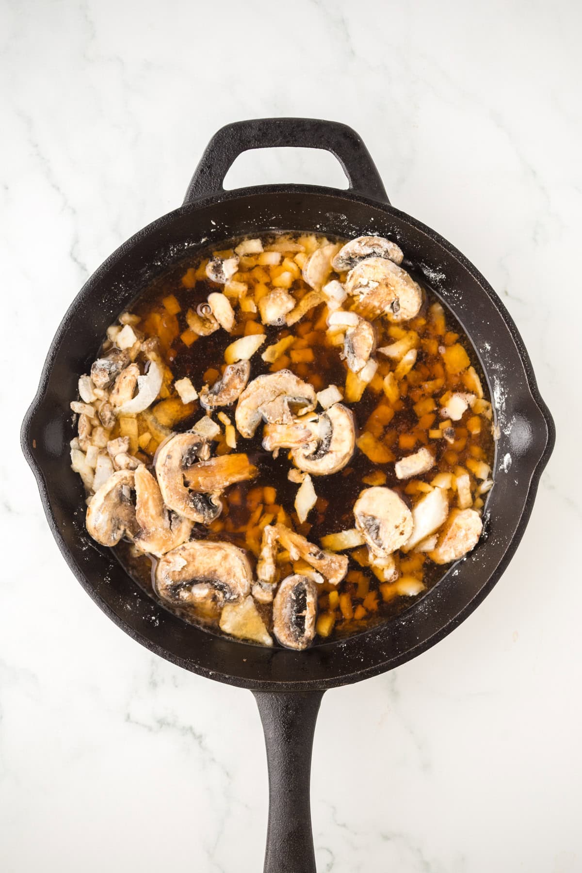 A black cast iron skillet containing chopped steak, sliced mushrooms, chopped onions, and a brown liquid, placed on a white marble surface.