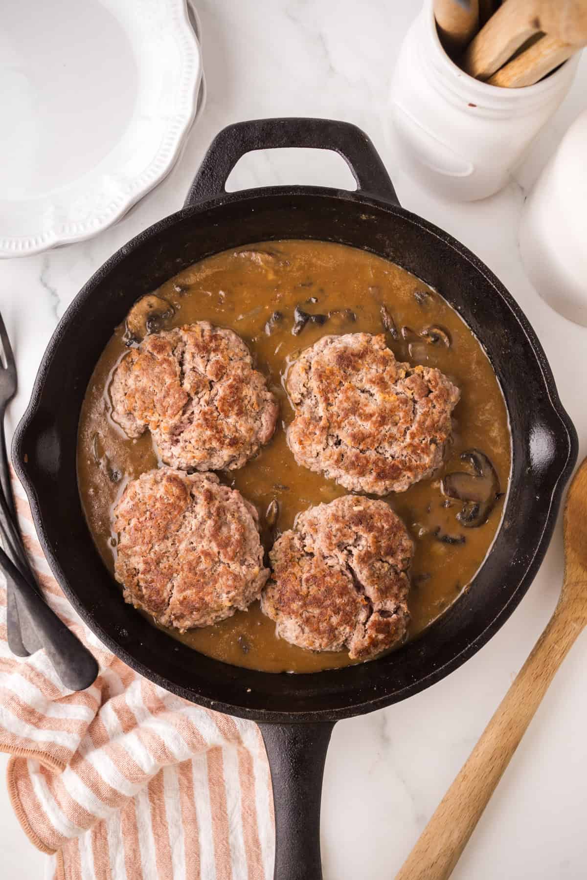 A cast iron skillet with four browned chopped steak patties in mushroom gravy sits on a white marble surface, accompanied by a striped towel, wooden spoon, and utensils.