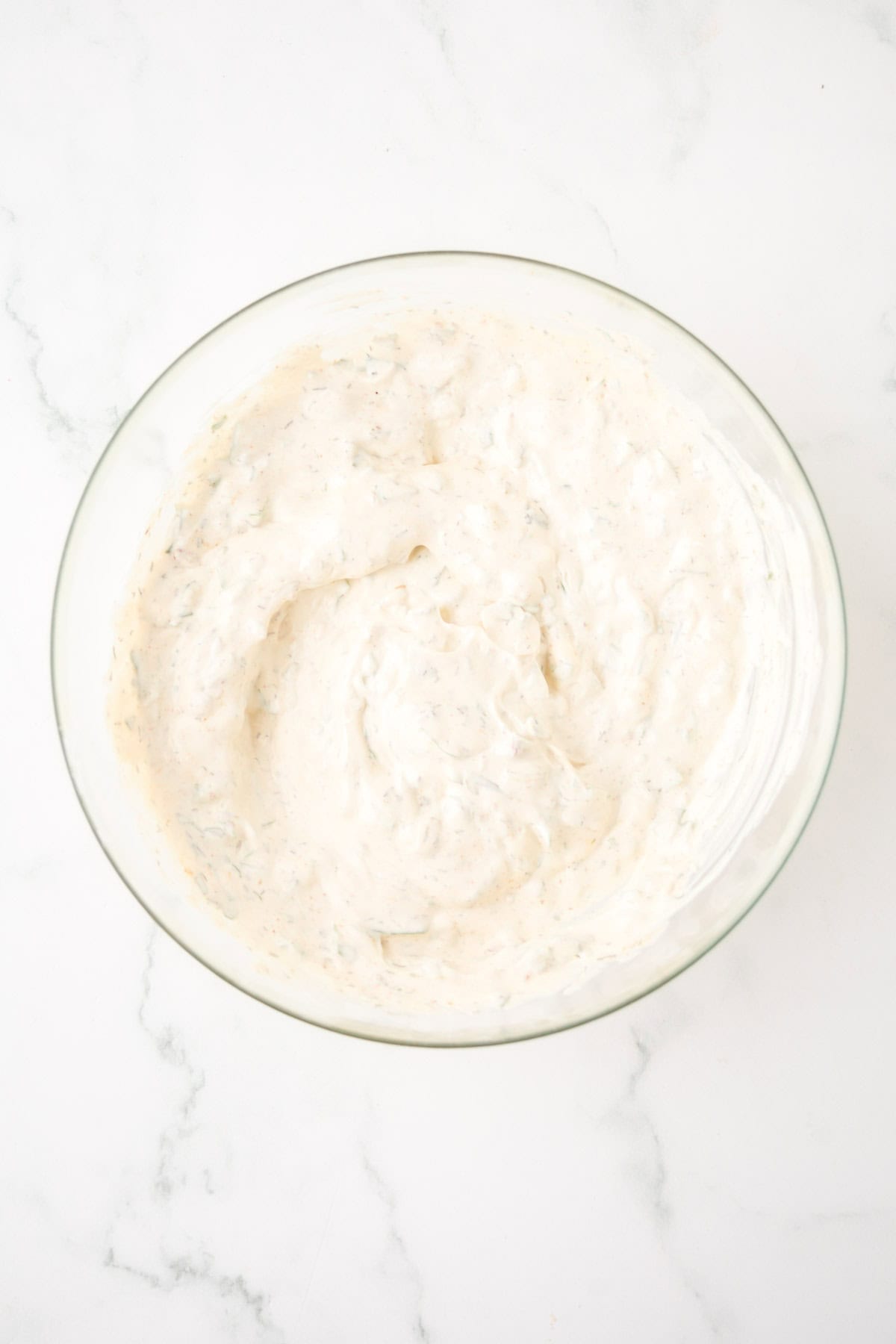 A glass bowl filled with creamy, white Knorr veggie spinach dip, flecked with herbs, sits on a white marble surface.