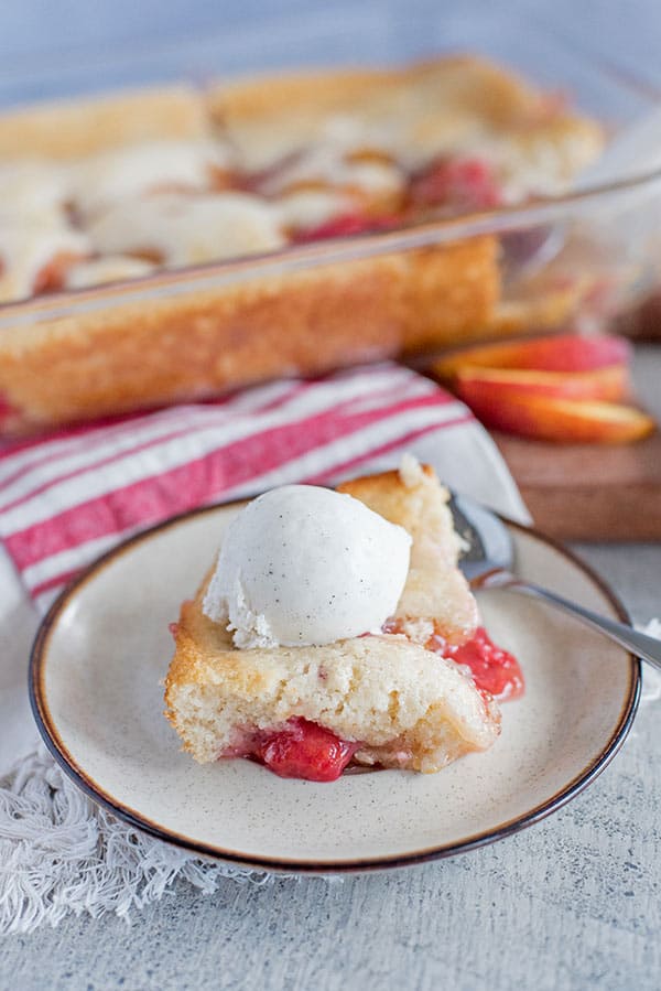 A slice of peach raspberry cobbler and strawberry cobbler topped with vanilla ice cream sits on a plate with a spoon. In the background, a glass baking dish with more cobbler and fresh peach slices are visible.