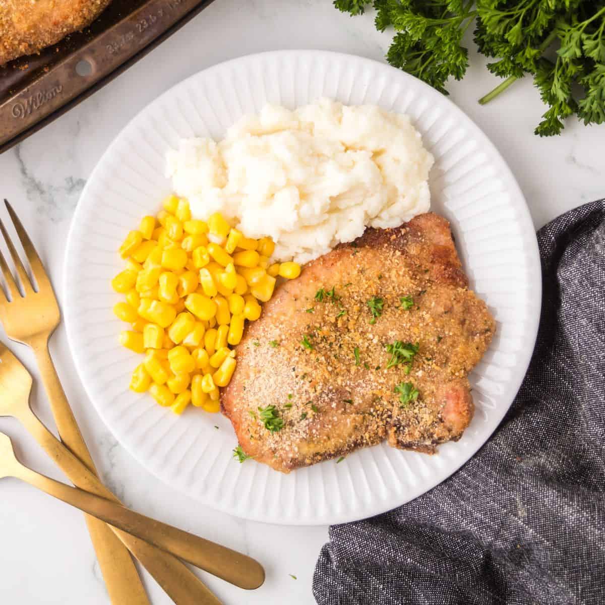 A white plate with breaded pork chop, mashed potatoes, and corn. Gold-colored utensils are placed to the left, with a bunch of parsley and a dark napkin nearby on a white surface.