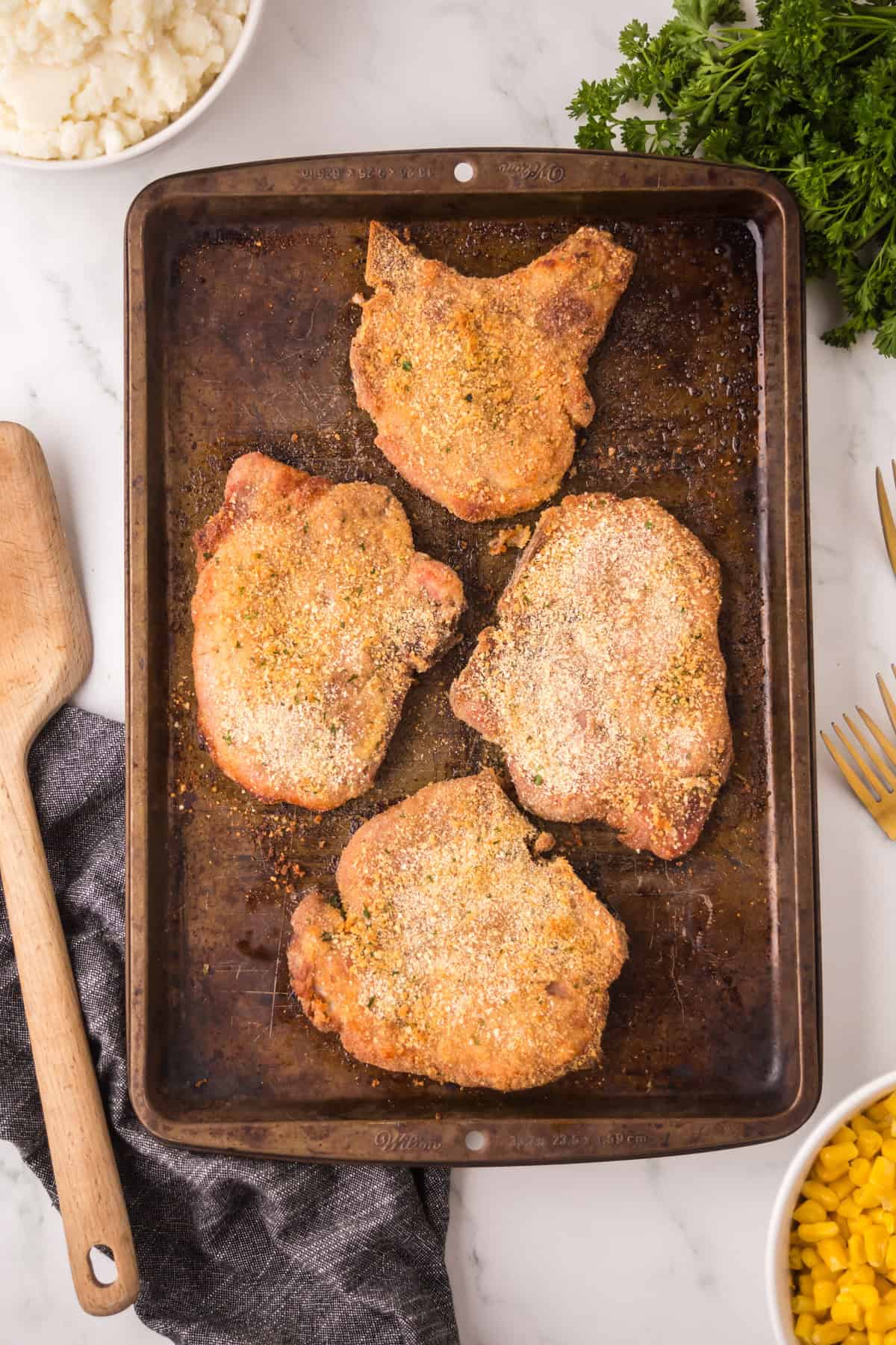 A baking sheet with four shake and bake pork chops, surrounded by a bowl of mashed potatoes, a bunch of parsley, a bowl of corn, a wooden spatula, and a gray napkin on a marble surface.