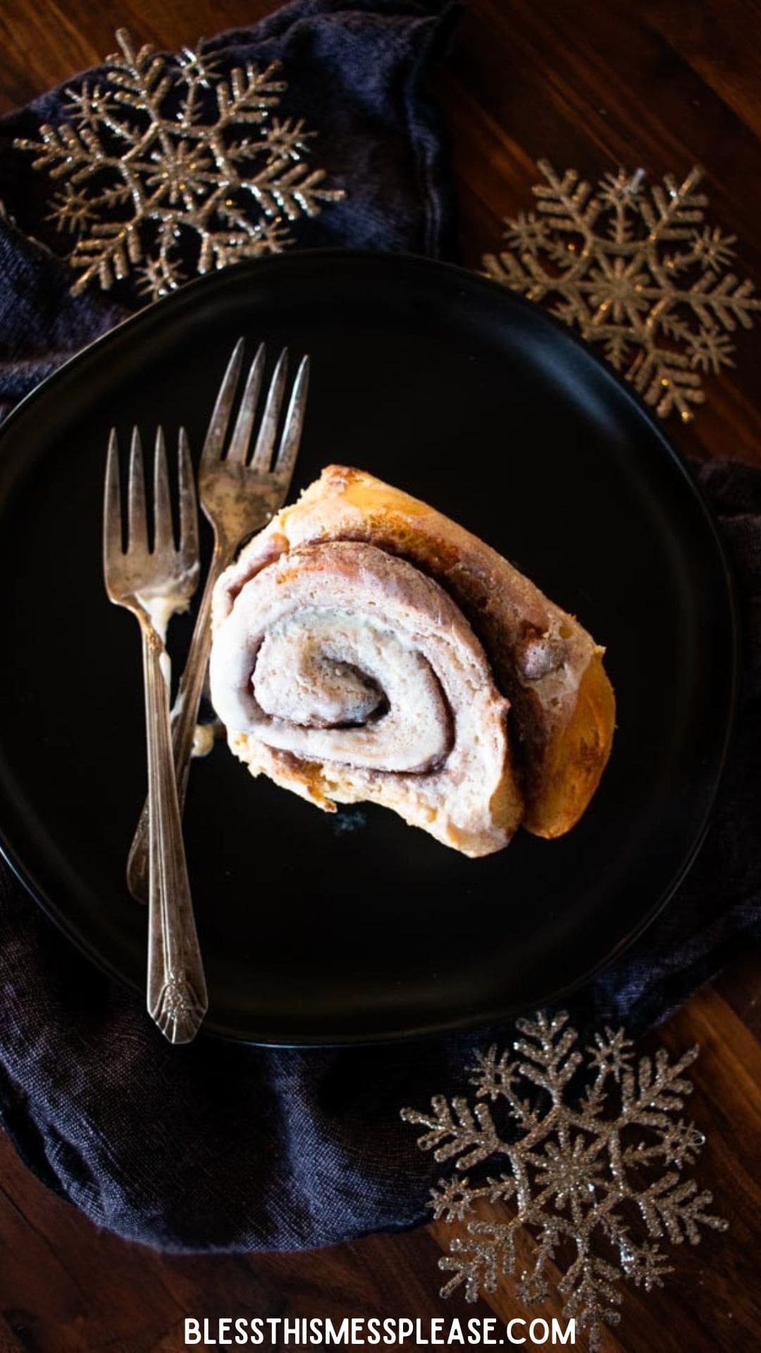 A frosted gingerbread cinnamon roll on a black plate with two forks, set on a dark cloth. Decorative gold snowflakes and the website are also visible.