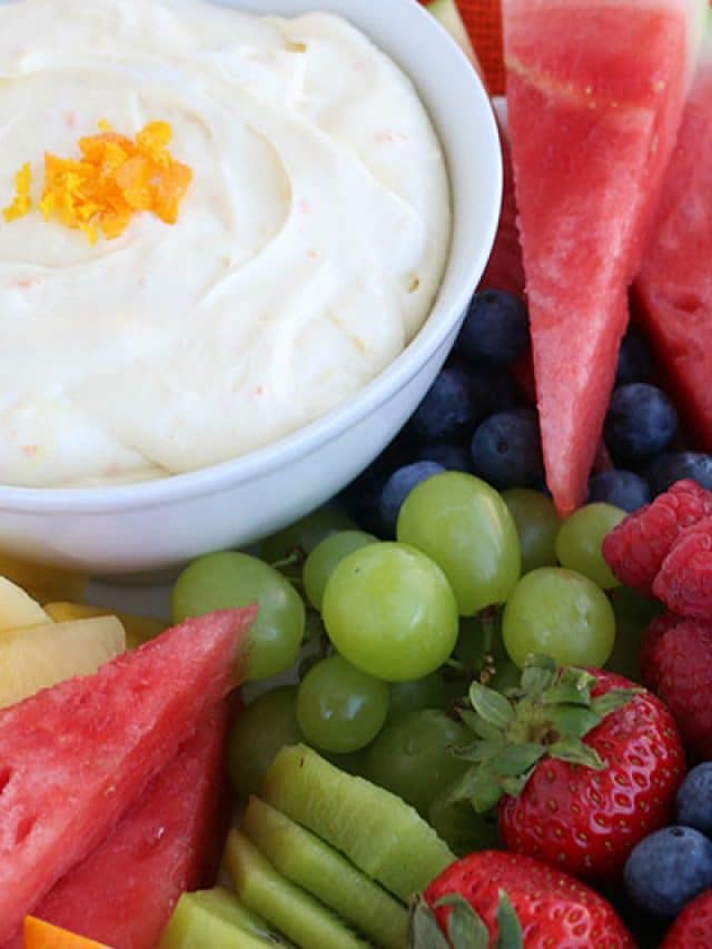 A bowl of creamy dip garnished with orange zest is surrounded by fresh fruit, including watermelon slices, green grapes, blueberries, raspberries, strawberries, kiwi, and pineapple.