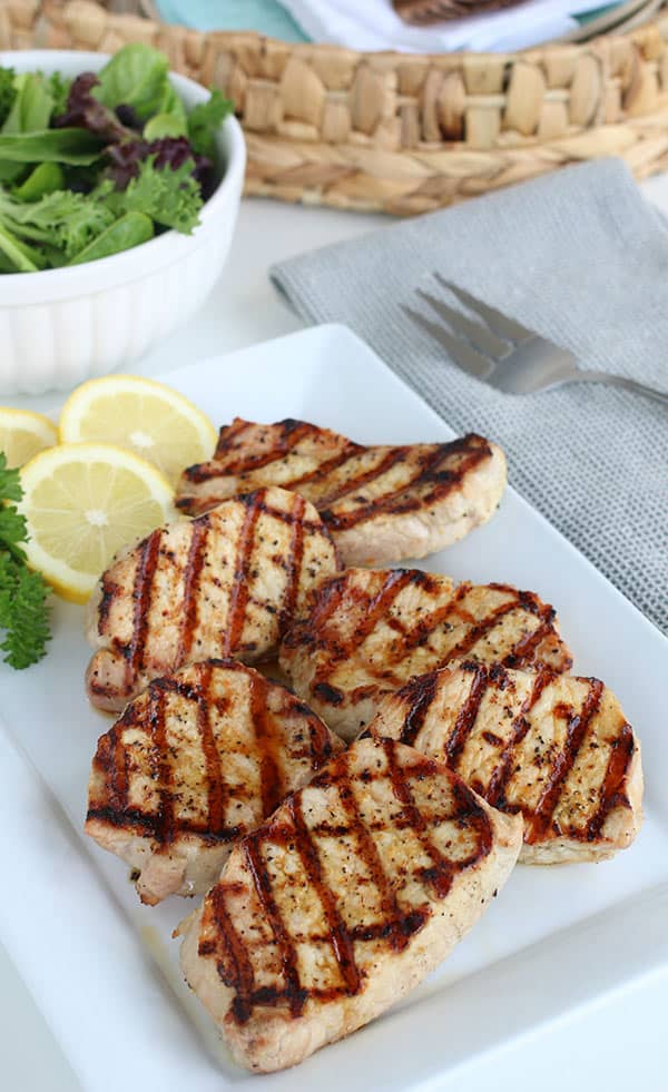 Lemon pepper porkchops with char marks on a white rectangular plate, garnished with lemon slices and parsley, alongside a bowl of mixed greens and a fork for a fresh, flavorful meal.