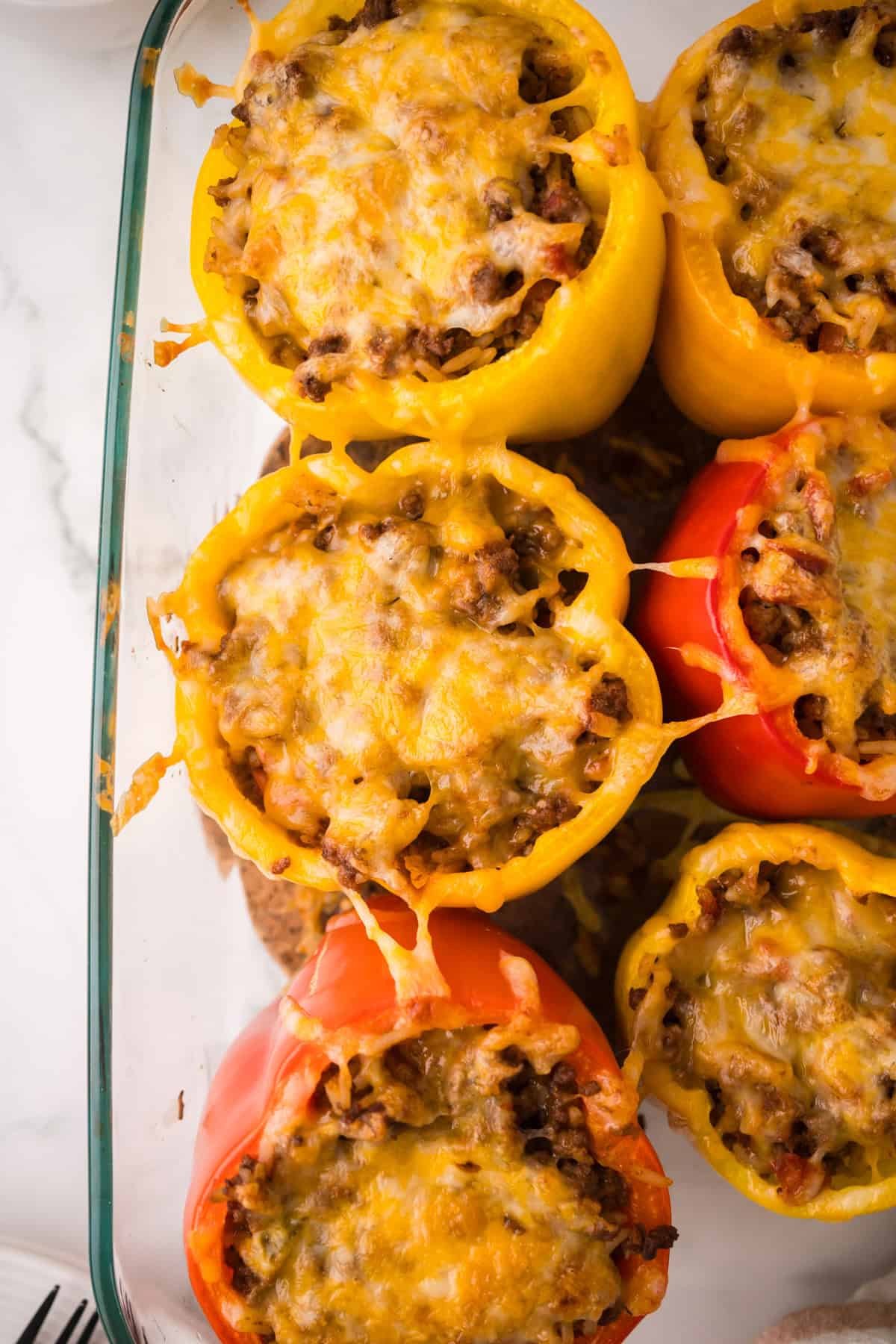 A glass baking dish filled with stuffed peppers with ground beef and melted cheese, featuring vibrant yellow and red bell peppers, viewed from above on a white surface featuring sides for what to serve with stuffed peppers.