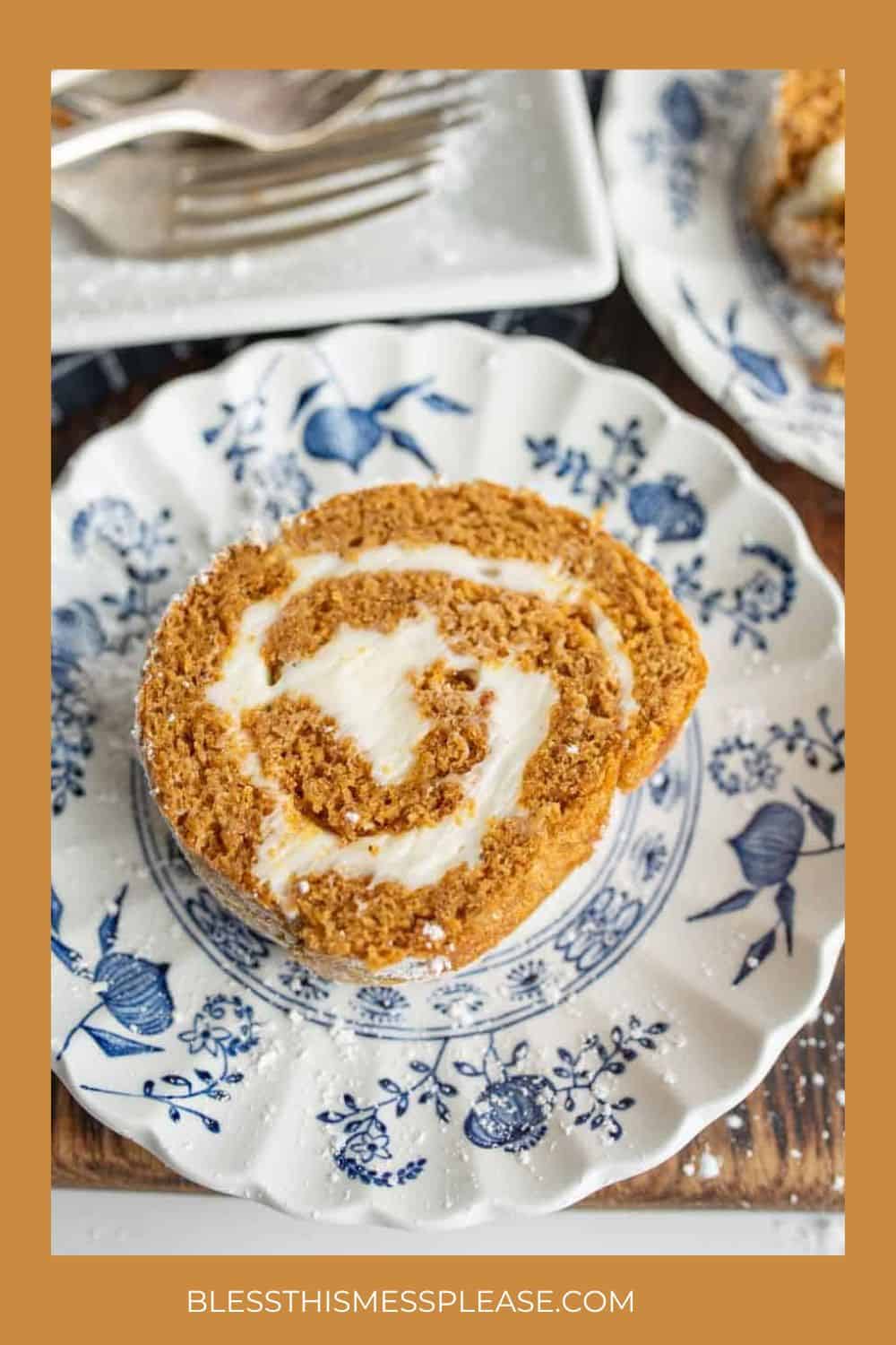 A slice of pumpkin roll cake with cream cheese filling, a classic among fall desserts, sits on a decorative blue and white plate; a fork is nearby. The plate rests on a wooden surface with another plate partly visible showing off cozy fall desserts.