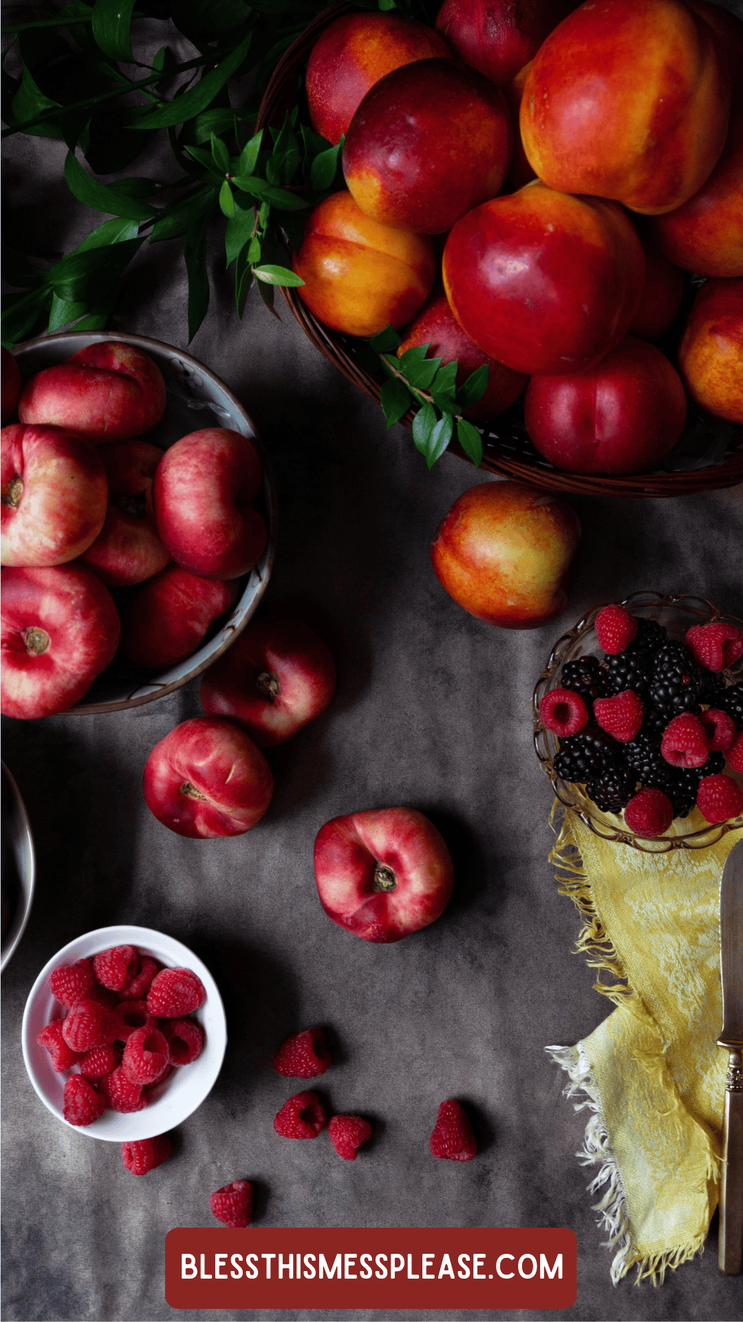 A flat lay of various fresh fruits, including nectarines, peaches, raspberries, and blackberries on a dark surface, with a yellow cloth and a label at the bottom—perfect inspiration for learning how to preserve plums.