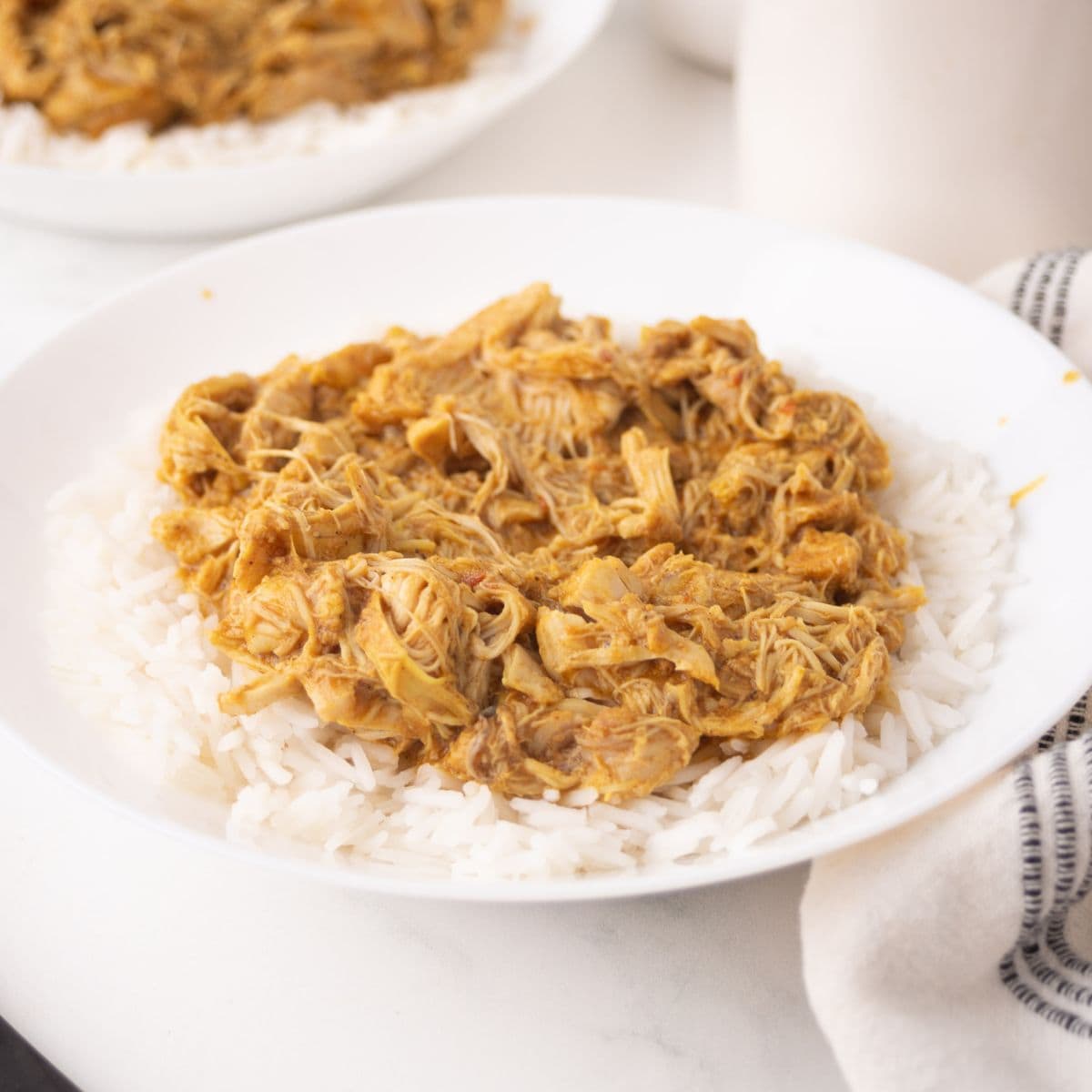 A white plate filled with shredded chicken korma in a creamy sauce served over a bed of white rice, with part of a striped napkin visible beside the plate.
