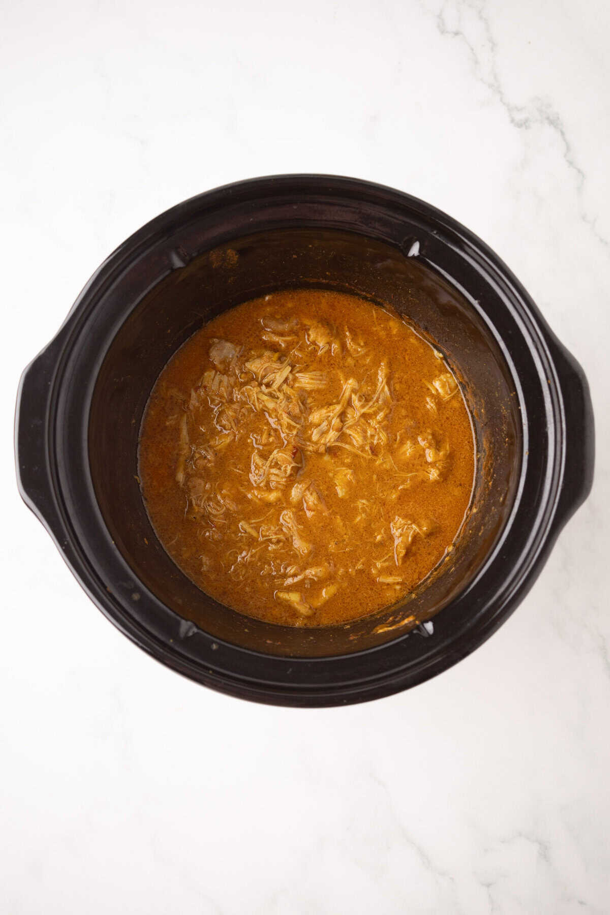 A crockpot filled with shredded chicken korma in a rich brown sauce, sitting on a white marble surface.