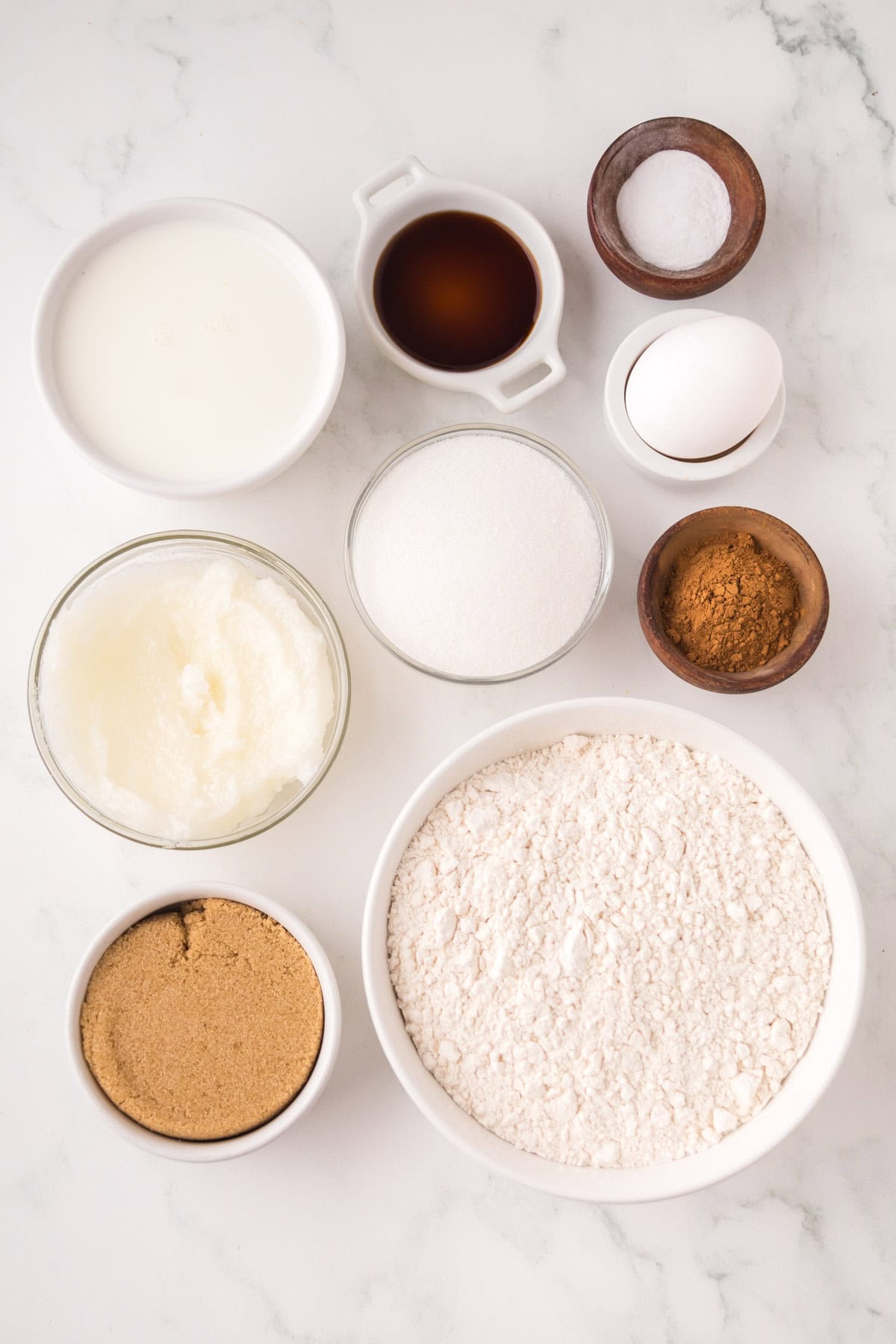 Assorted baking ingredients arranged on a white marble surface, including flour, sugar, brown sugar, shortening, cocoa powder, salt, an egg, milk, and vanilla extract in small bowls and dishes.