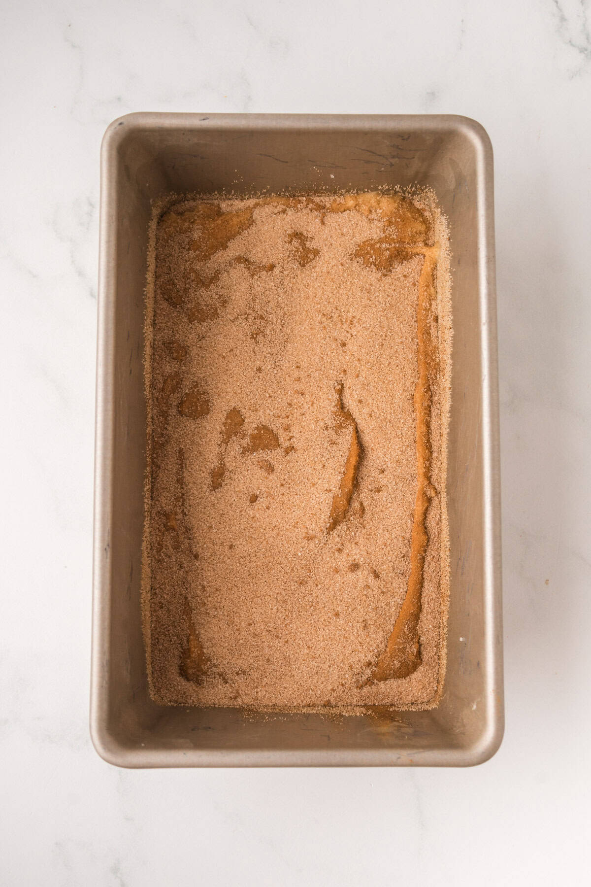 A metal loaf pan filled with a layer of cinnamon sugar mixture sits on a white marble surface.