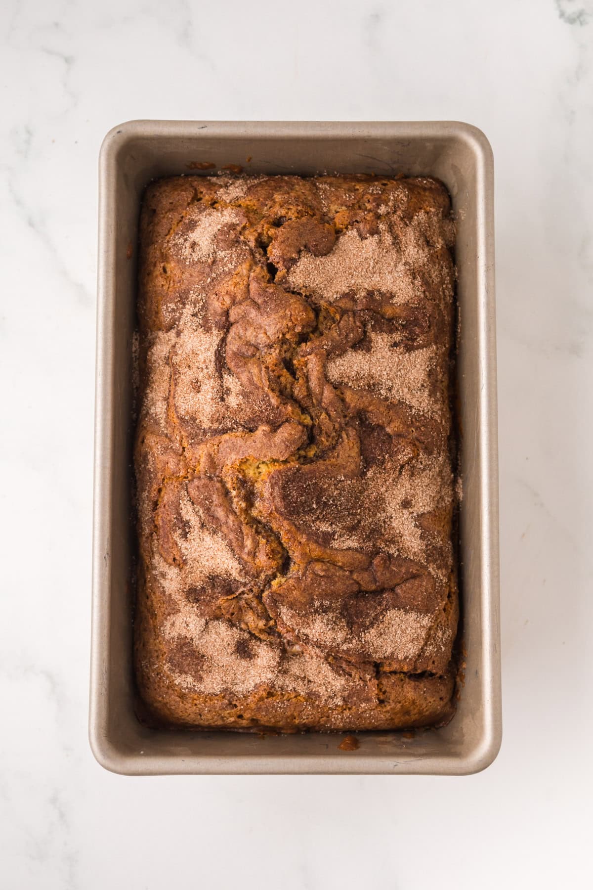 A loaf of baked bread with a cinnamon sugar topping sits in a metal baking pan on a white marble surface.