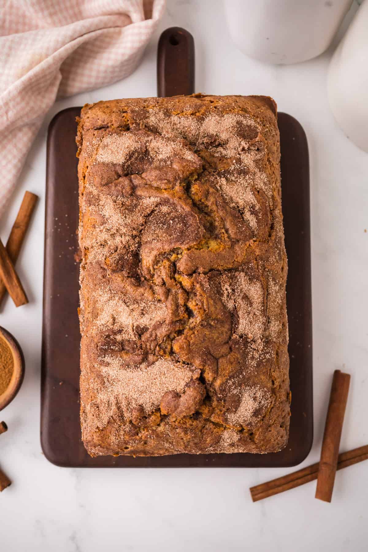 A loaf of cinnamon sugar bread sits on a dark wooden cutting board, surrounded by cinnamon sticks and a pink checkered cloth on a white marble surface. The bread has a swirled, sugar-dusted top.