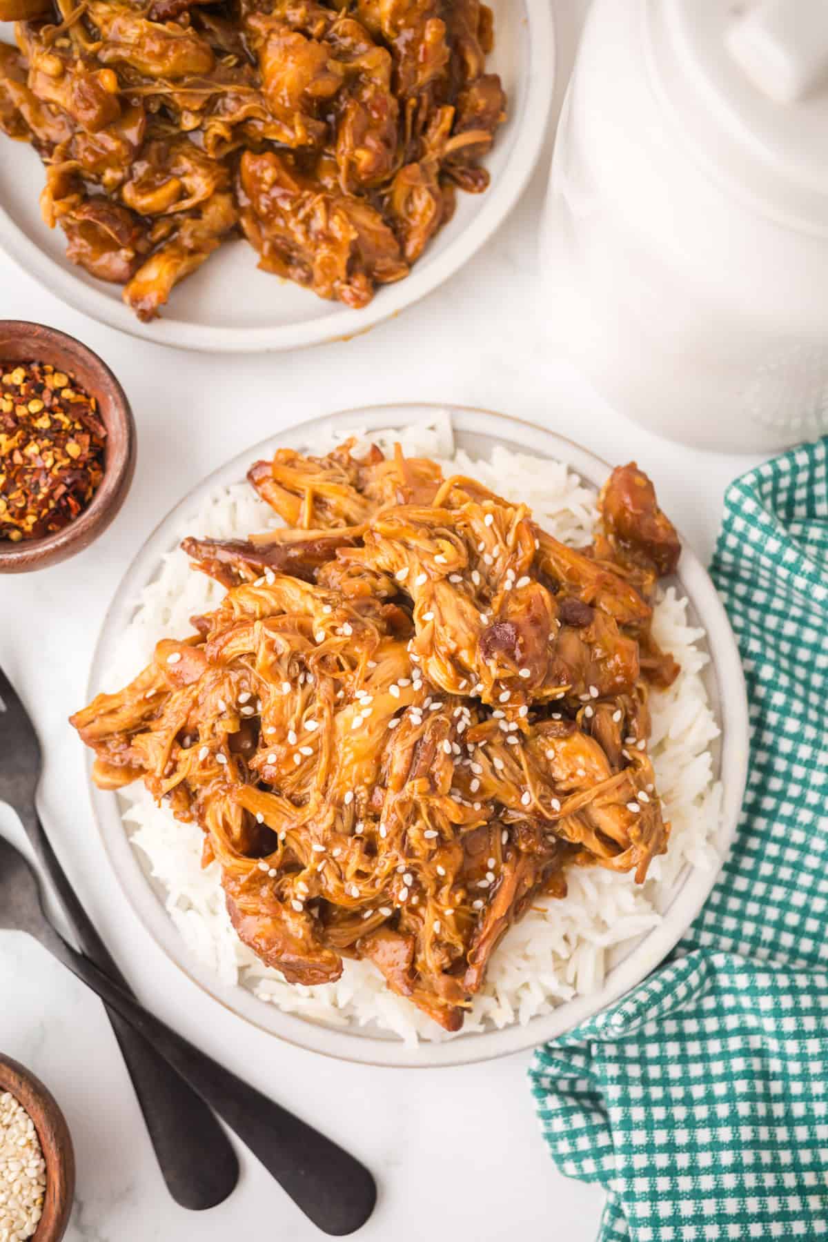 A plate of crockpot garlic honey chicken, shredded and saucy, served over white rice and topped with sesame seeds. A green checkered cloth, utensils, and small bowls of spices sit nearby, with a second plate in the background.
