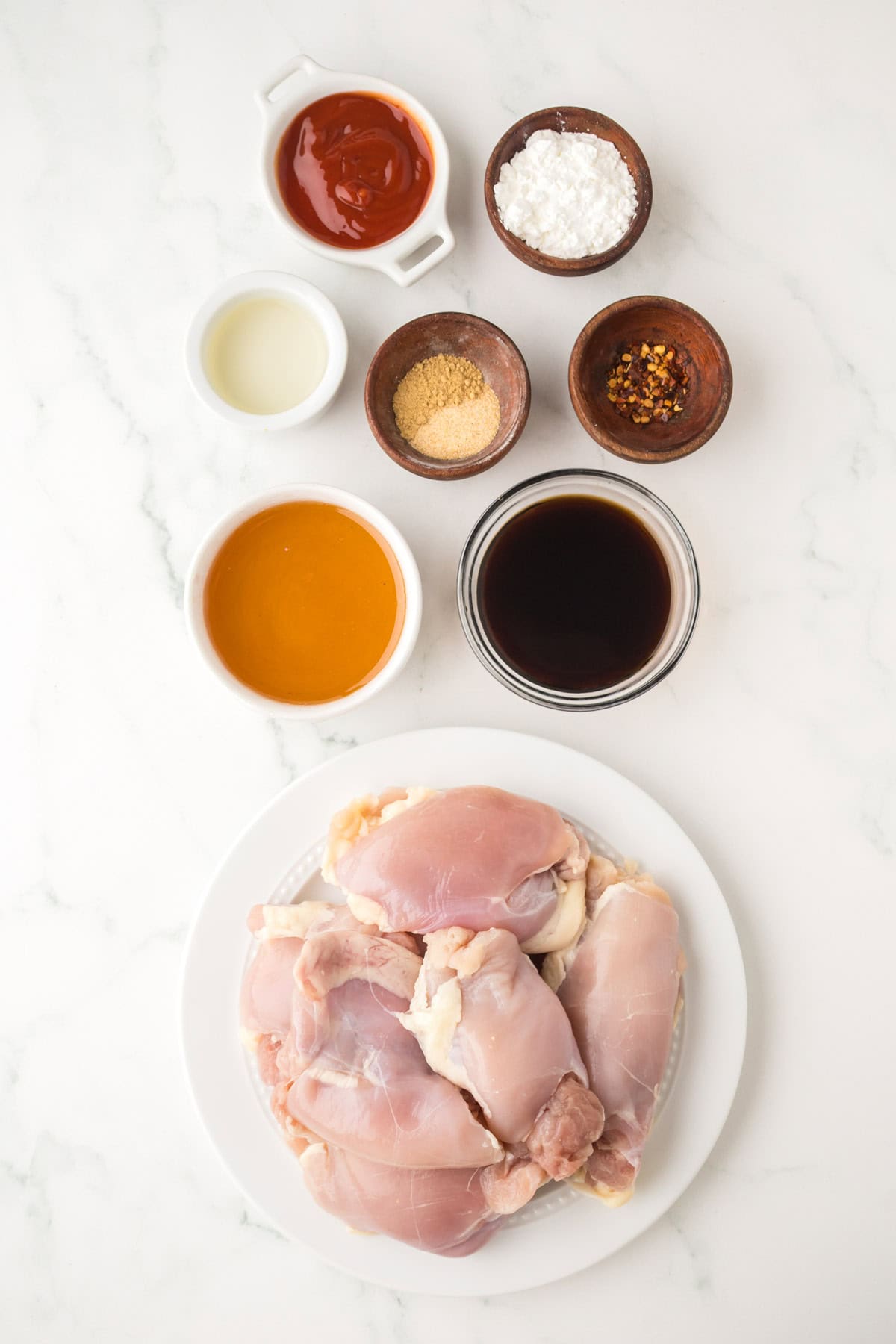 A plate of raw chicken thighs with small bowls of soy sauce, honey, ketchup, cornstarch, oil, garlic powder, and crushed red pepper flakes—perfect for making crockpot garlic honey chicken—on a white marble surface.