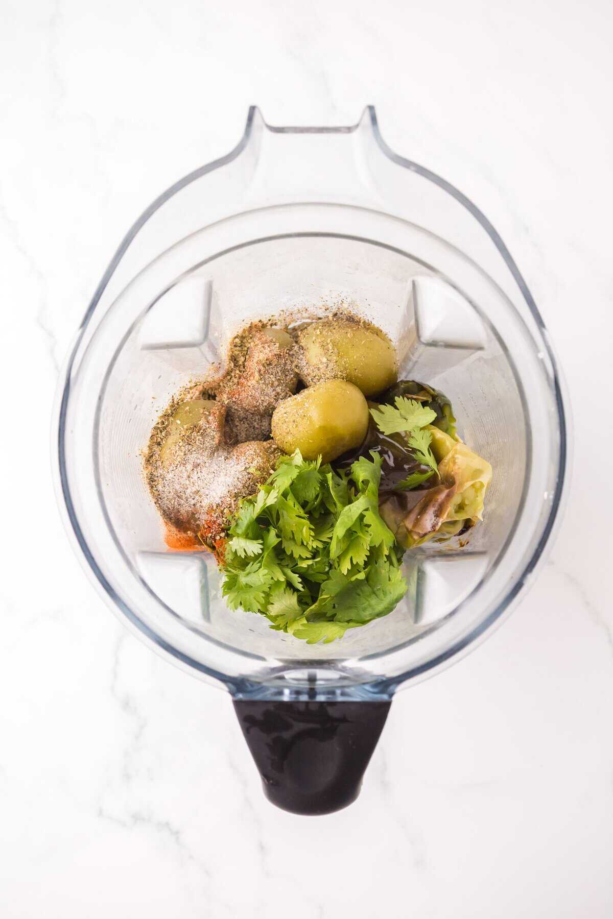 A top-down view of a blender containing green olives, fresh cilantro, spices, and other ingredients for pork chili verde on a white marble surface, ready to be blended.