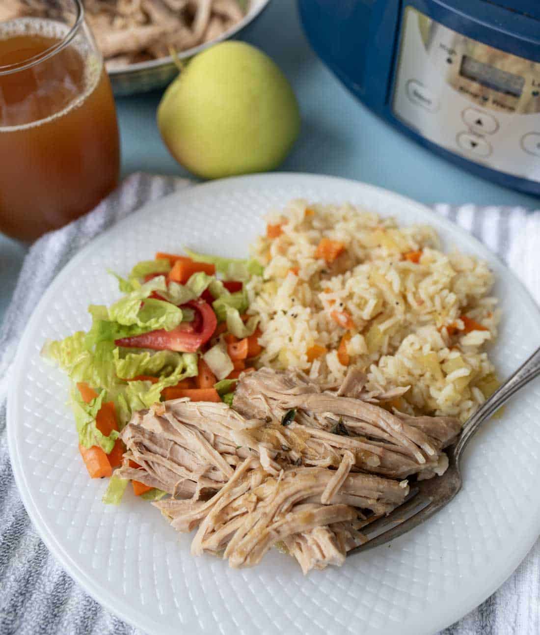 A white plate with shredded pork, vegetable rice, and a lettuce salad. A fork is on the plate. In the background are a glass of iced tea, a pear, a bowl of shredded pork, and a pressure cooker.