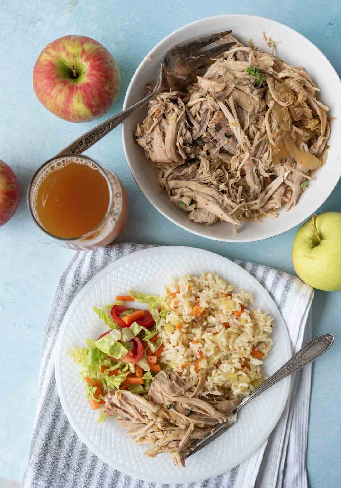 A plate with shredded pork, rice with vegetables, and a small salad sits on a striped towel. Nearby are a bowl of shredded pork, apples, and a glass of apple juice on a light blue surface.