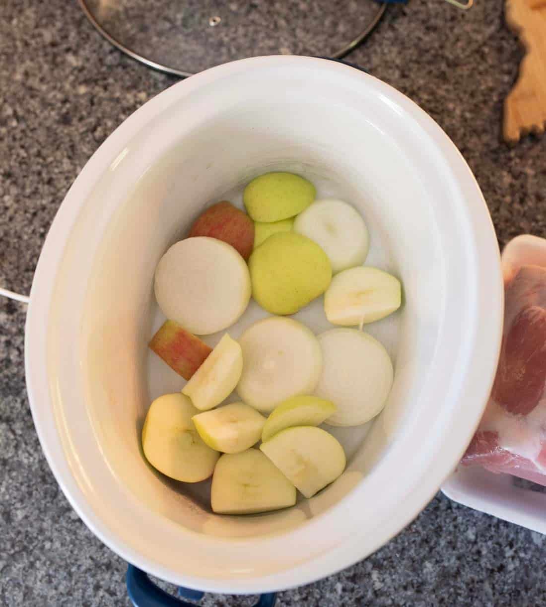 A slow cooker filled with slices of apple and onion placed on a gray countertop, with a tray of raw meat partially visible to the right.