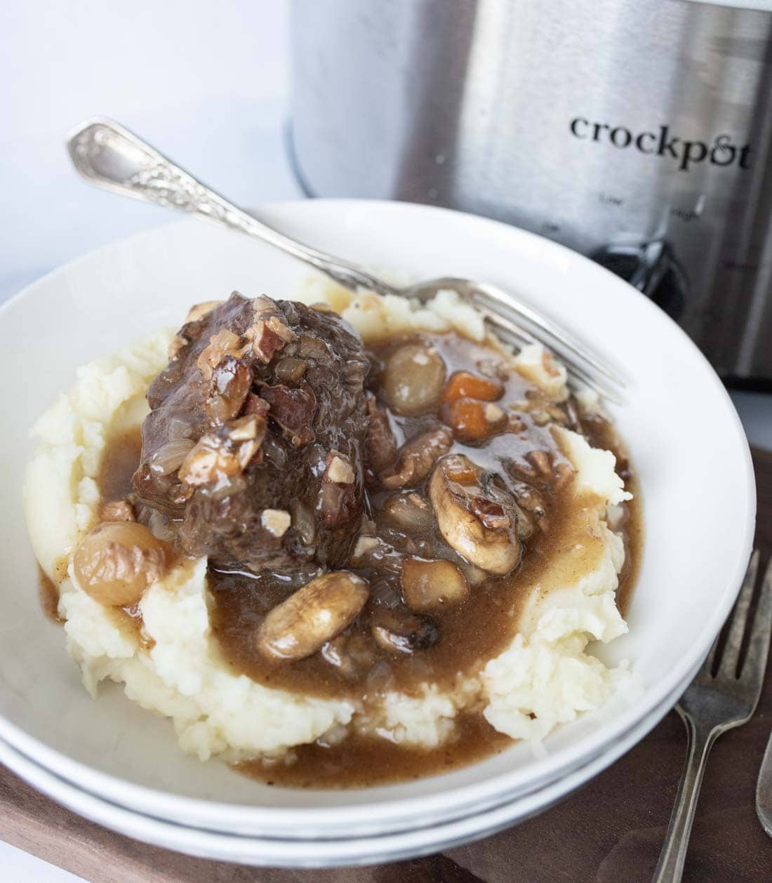 A white bowl filled with mashed potatoes topped with beef stew containing onions, mushrooms, and carrots, with a fork and a crockpot in the background.