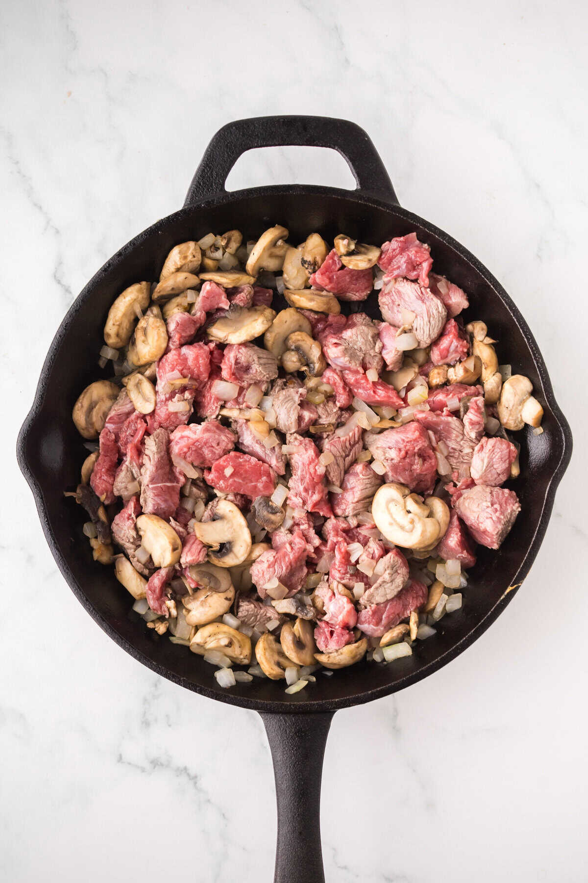 A cast iron skillet filled with sliced beef, mushrooms, and chopped onions being sautéed on a white marble surface—perfect ingredients for a classic slow cooker beef stroganoff.