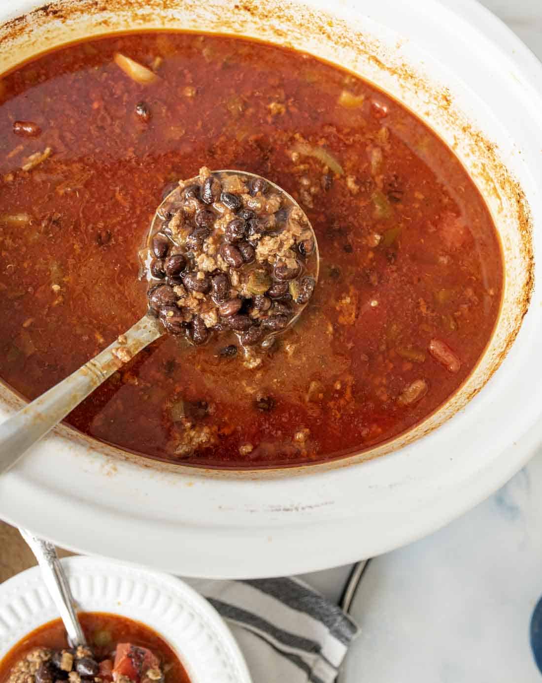 A ladle scoops up hearty chili filled with ground meat, black beans, and vegetables from a white slow cooker. A bowl of chili sits nearby on a striped napkin.