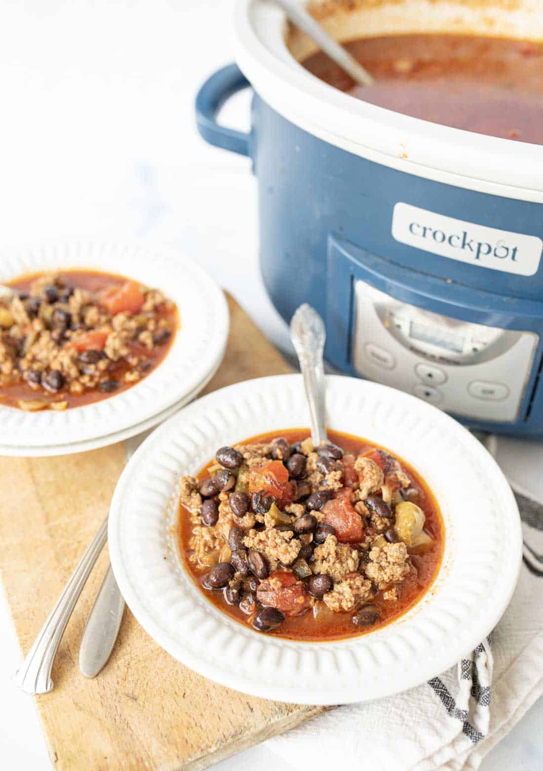 Two bowls of chili with ground beef, black beans, and tomatoes sit on a wooden board next to a blue Crockpot filled with more chili. Spoons rest in the bowls and by the Crockpot.