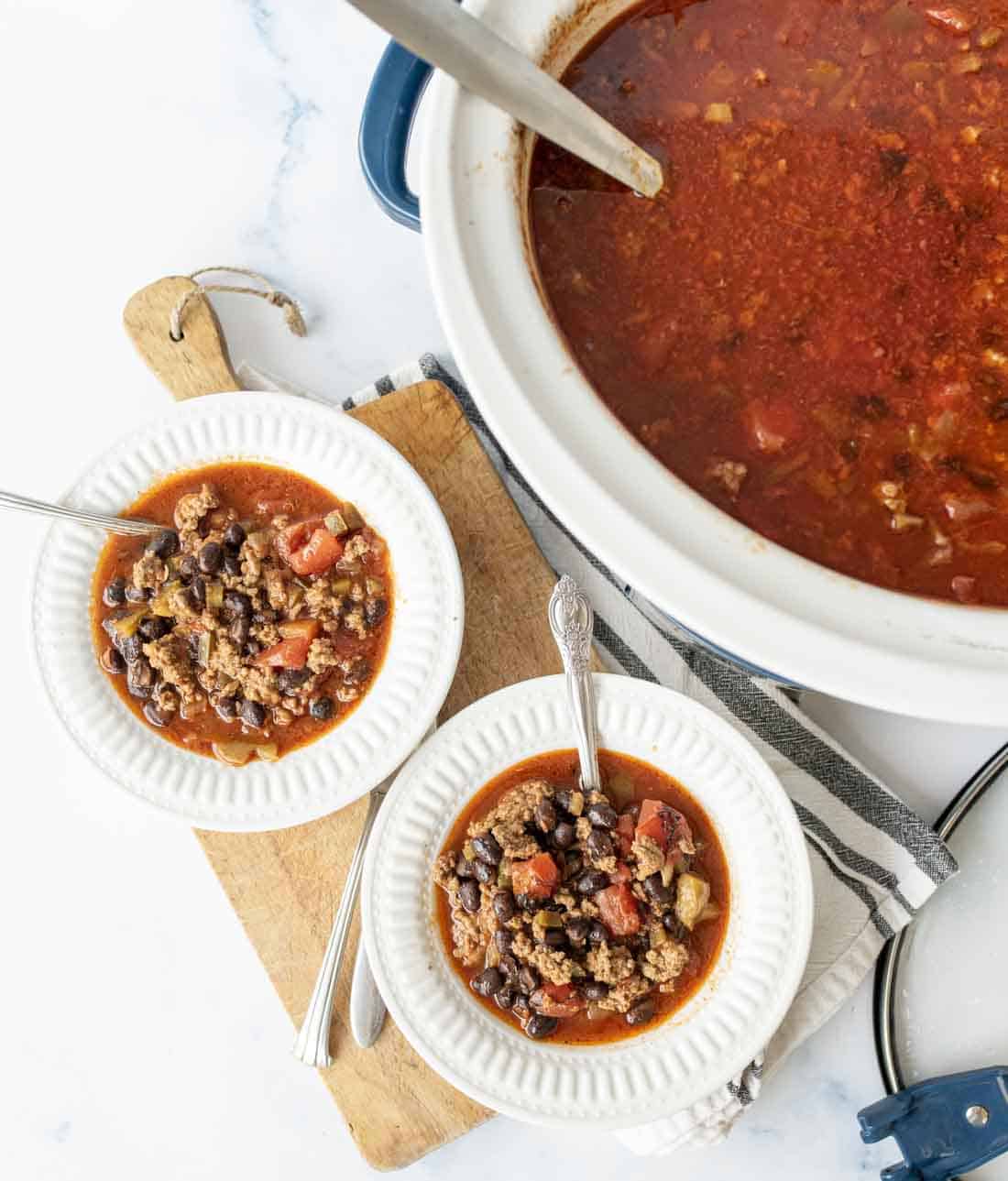 A white crockpot filled with chili sits on a counter next to two white bowls of chili with black beans, tomatoes, and ground meat, each with a spoon, all placed on a wooden board with a striped towel underneath.