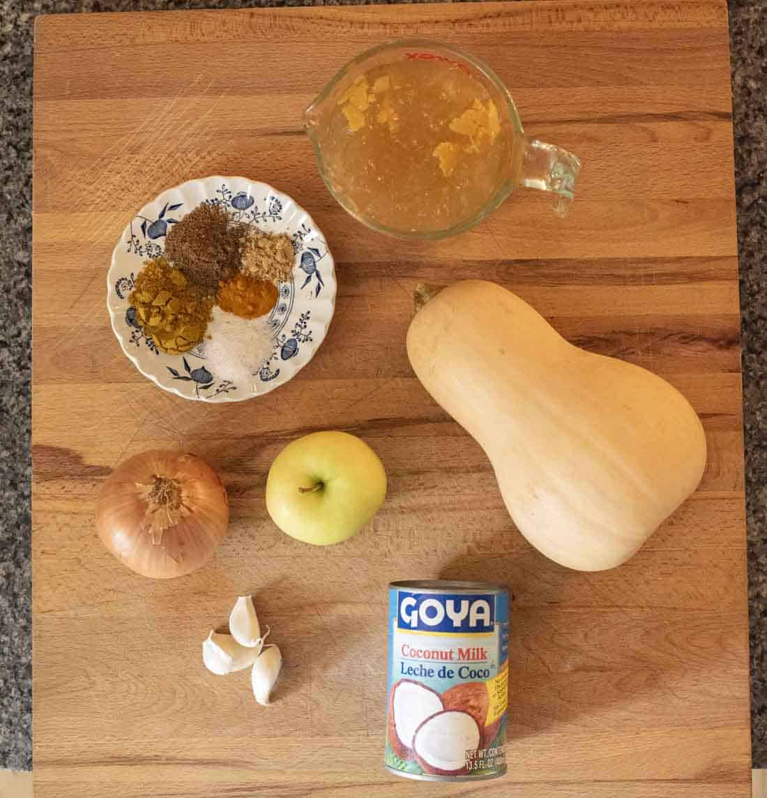 Ingredients for a recipe displayed on a wooden surface: a measuring cup with liquid, a plate with spices, a butternut squash, an onion, a green apple, garlic cloves, and a can of Goya coconut milk.