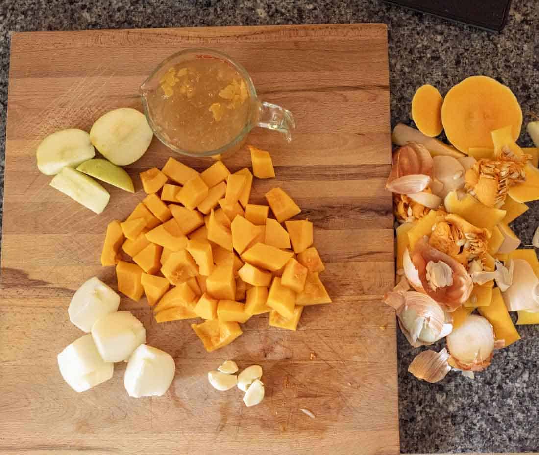 A wooden cutting board with chopped butternut squash, apple slices, peeled onions, garlic cloves, a measuring cup of broth, and a pile of vegetable peels on the side, set on a granite countertop.
